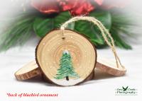 A hand-painted evergreen tree in the snow on the back of an eastern bluebird wood slice ornament at covecreekphotography.com.