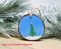 A hand-painted evergreen tree in the snow on the back of a chickadee wood slice Christmas ornament.