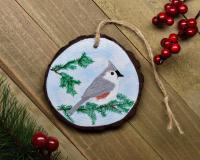 A handmade wood slice ornament with a hand-painted gray bird perched on an evergreen limb at Cove Creek Photography.