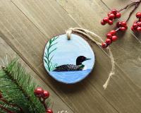 A hand-painted common loon on a pond wood slice Christmas ornament is displayed on a wooden board at Cove Creek Photography.