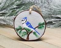 A hand-painted wood slice Christmas ornament with a blue jay perched in the top of a cedar tree with a winter gray background at Cove Creek Photography.