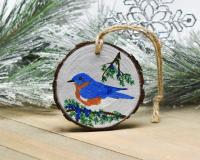 A hand-painted blue jay amid cedar branches and blue berries on a wood slice Christmas ornament at Cove Creek Photography.