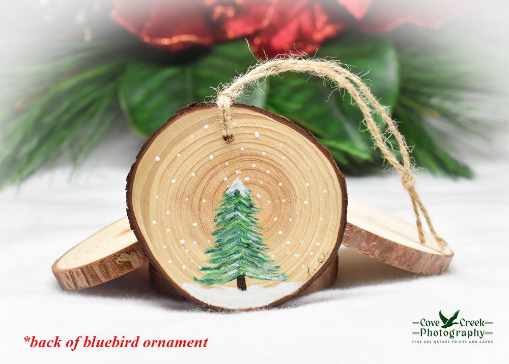 A hand-painted evergreen tree in the snow on the back of an eastern bluebird wood slice ornament at covecreekphotography.com.