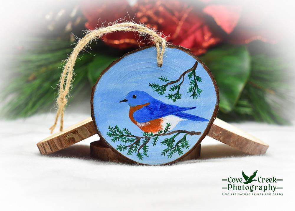 A hand-painted wood slice ornament with an eastern bluebird on a cedar branch.  Ornament is available at Cove Creek Photography.