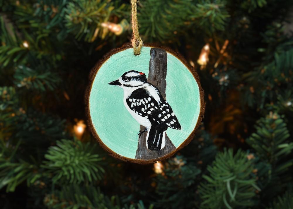 A wood slice Christmas ornament with a hand-painted downy woodpecker that is available for purchase at covecreekphotography.com.