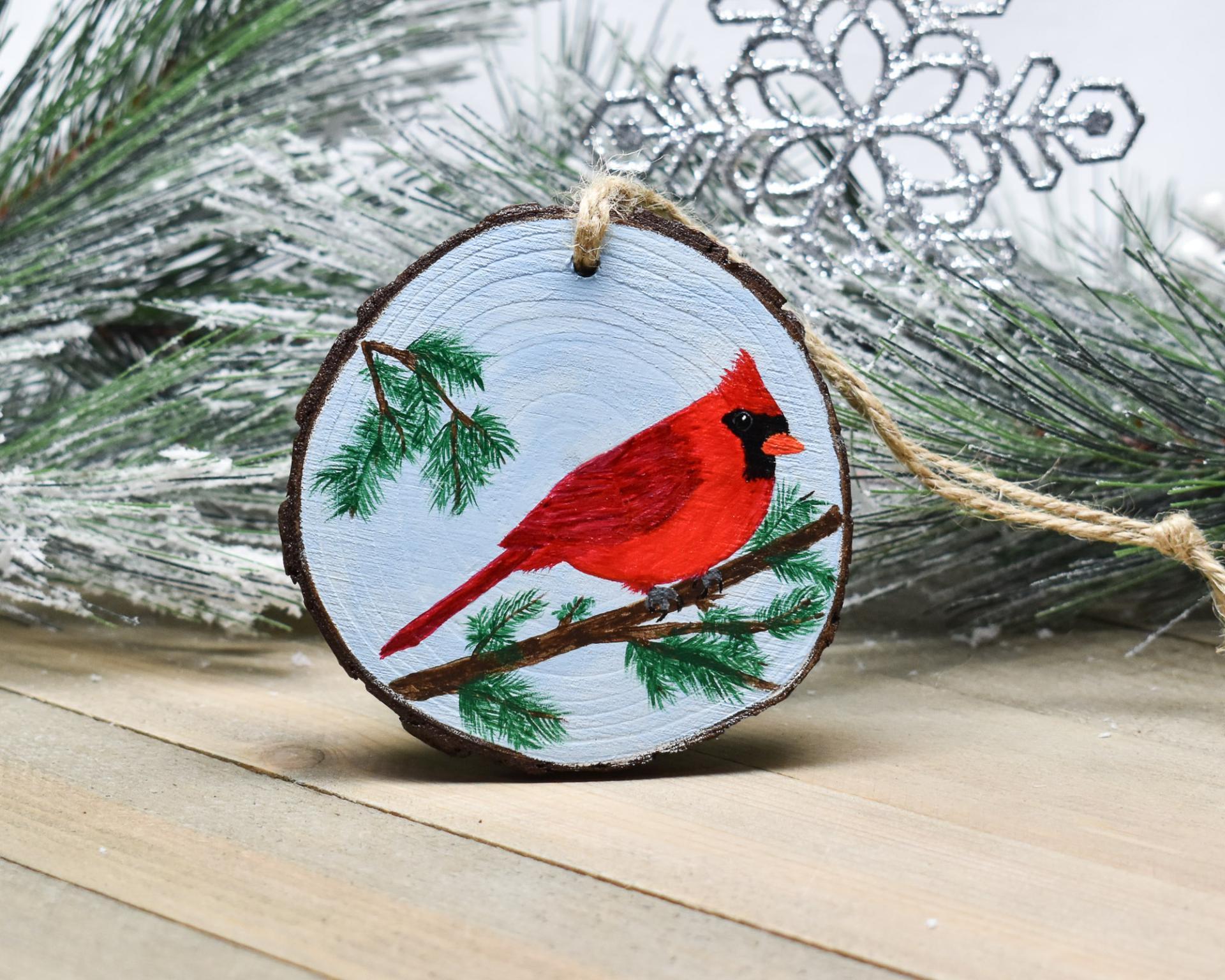 A hand-painted male northern cardinal perched on a cedar branch wood slice Christmas ornament at covecreekphotography.com.