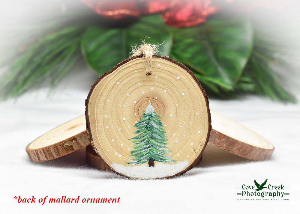 A hand-painted evergreen tree in snow on the back of a mallard wood slice ornament at covecreekphotography.com.