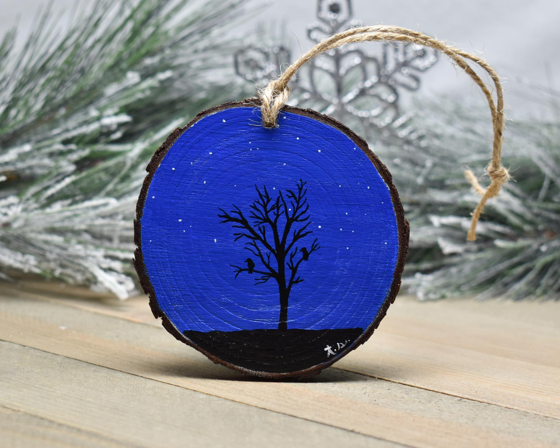 A dark blue night sky with stars and a silhouette of birds perching in a tree that has been hand-painted on the back of a white-breasted nuthatch wood slice Christmas ornament at covecreekphotography.com.