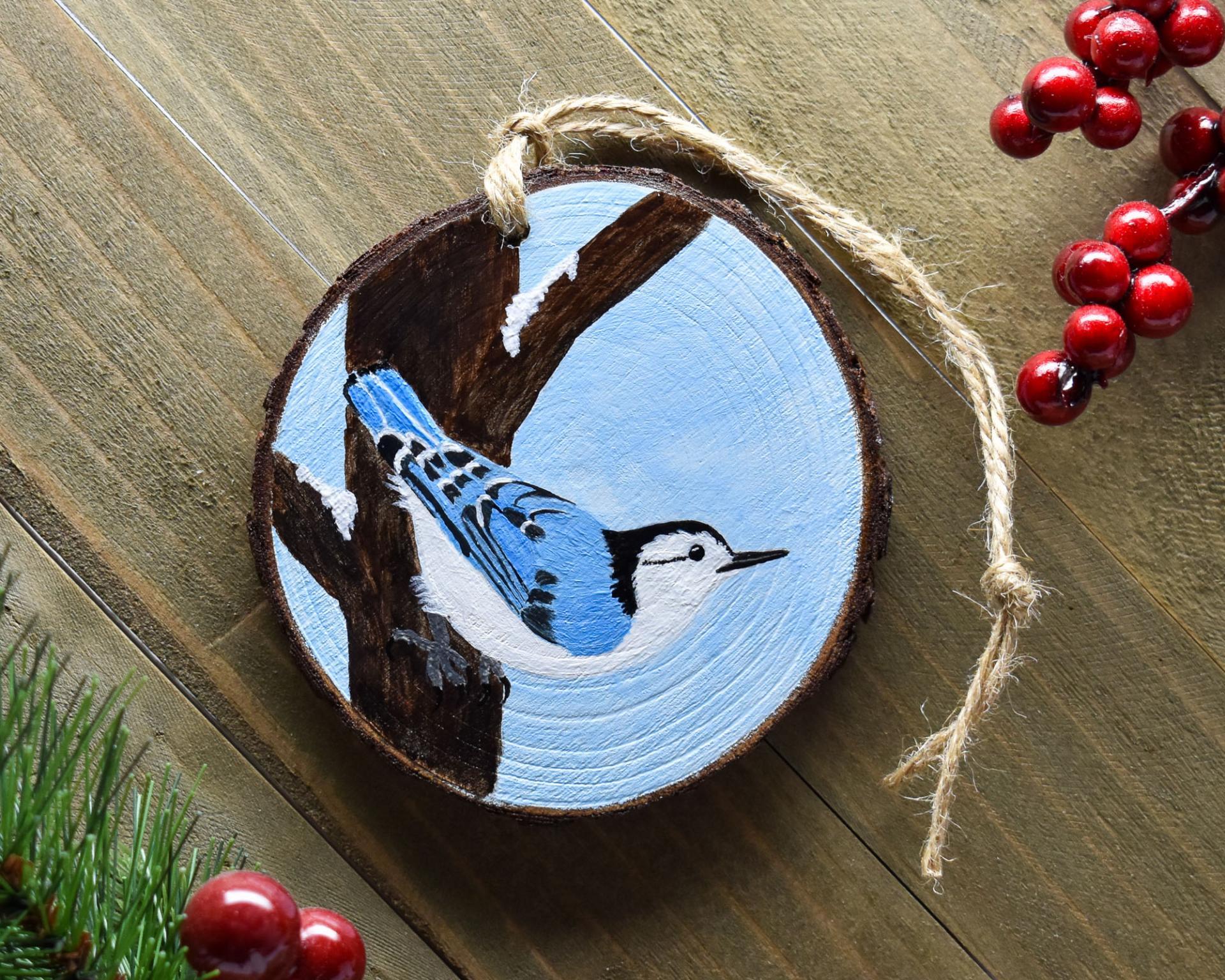 A hand-painted white-breasted nuthatch wood slice Christmas ornament displayed on weathered wood at Cove Creek Photography.