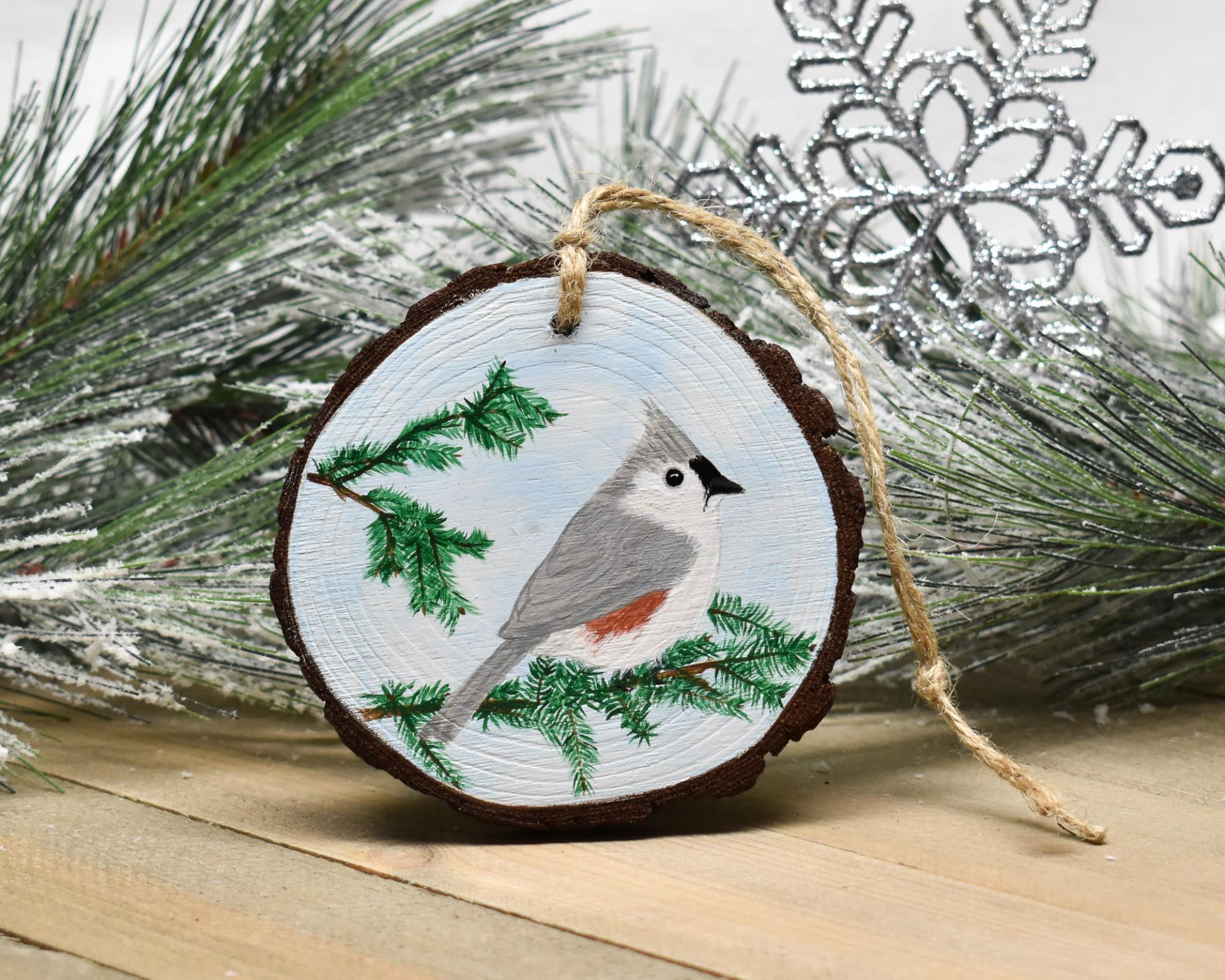 A tufted titmouse perched on an evergreen branch is hand-painted on a wood slice ornament at Cove Creek Photography.