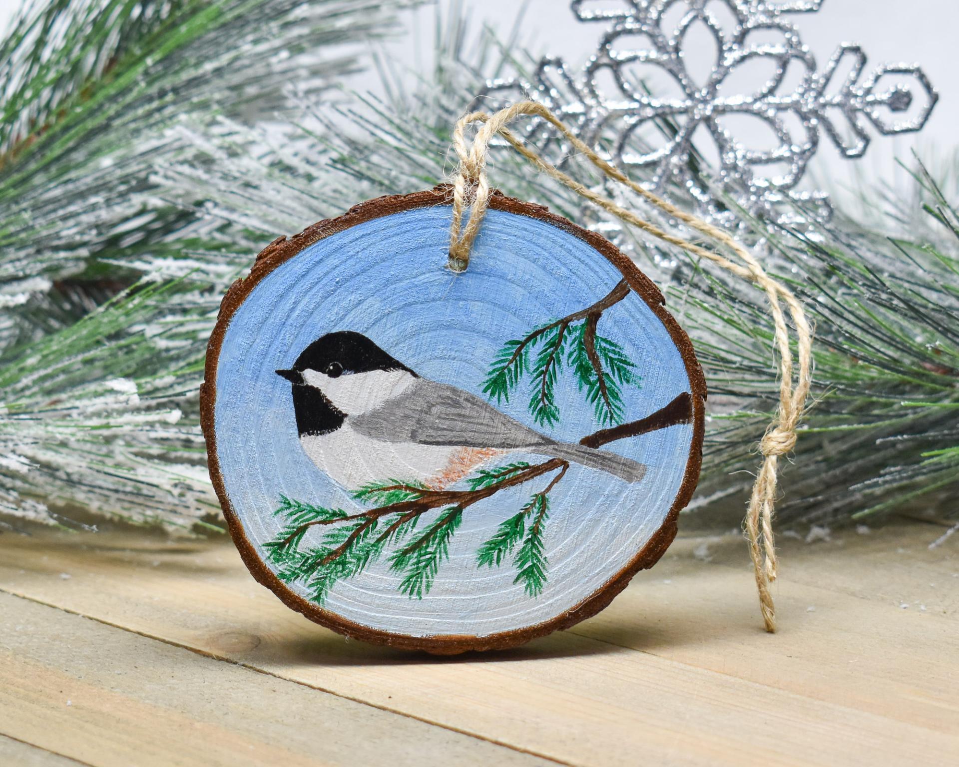 A Carolina chickadee perched on an evergreen limb is hand-painted on a wood slice Christmas ornament at Cove Creek Photography.
