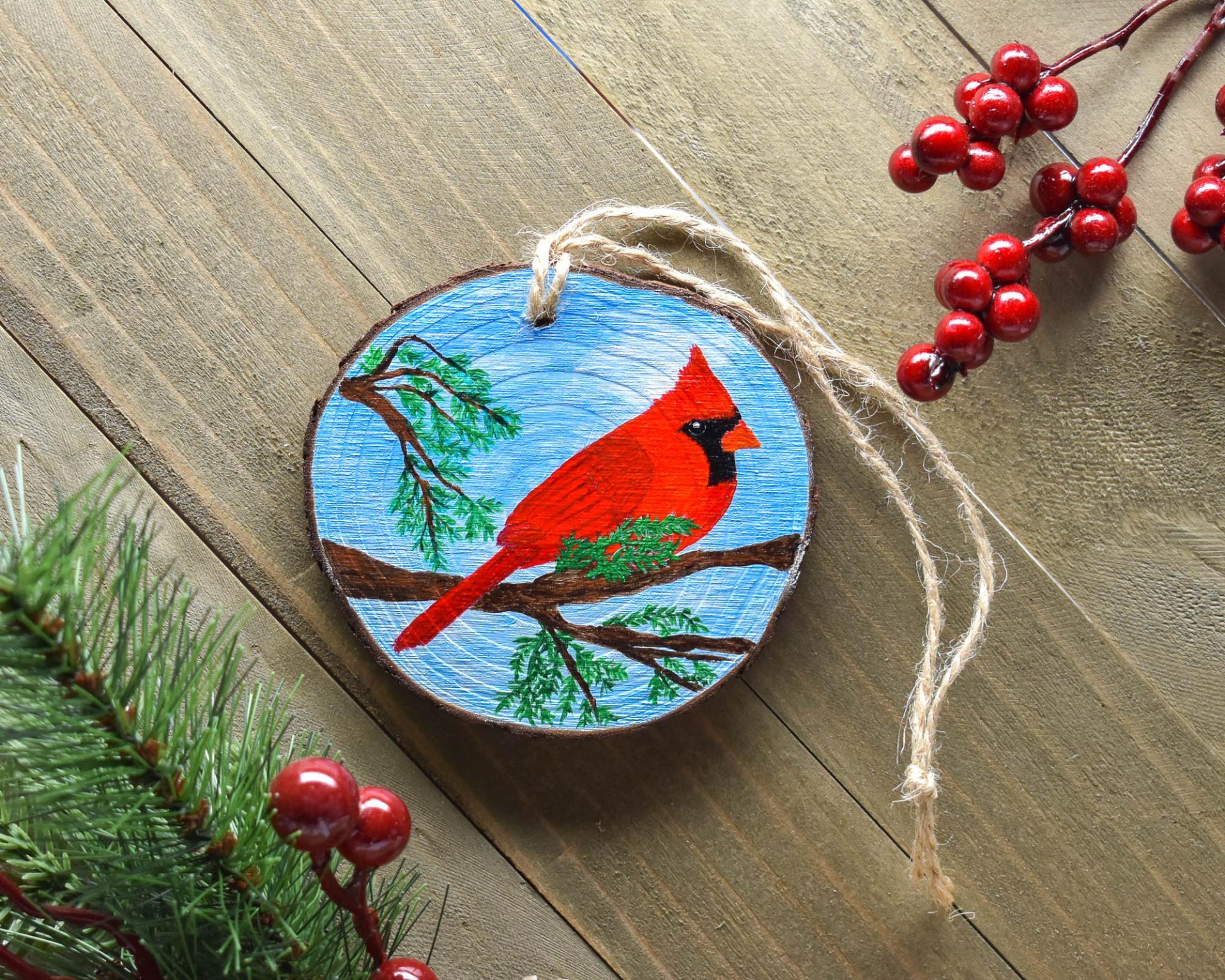 A hand-painted northern cardinal on a wood slice ornament is displayed on a rustic board background at Cove Creek Photography.