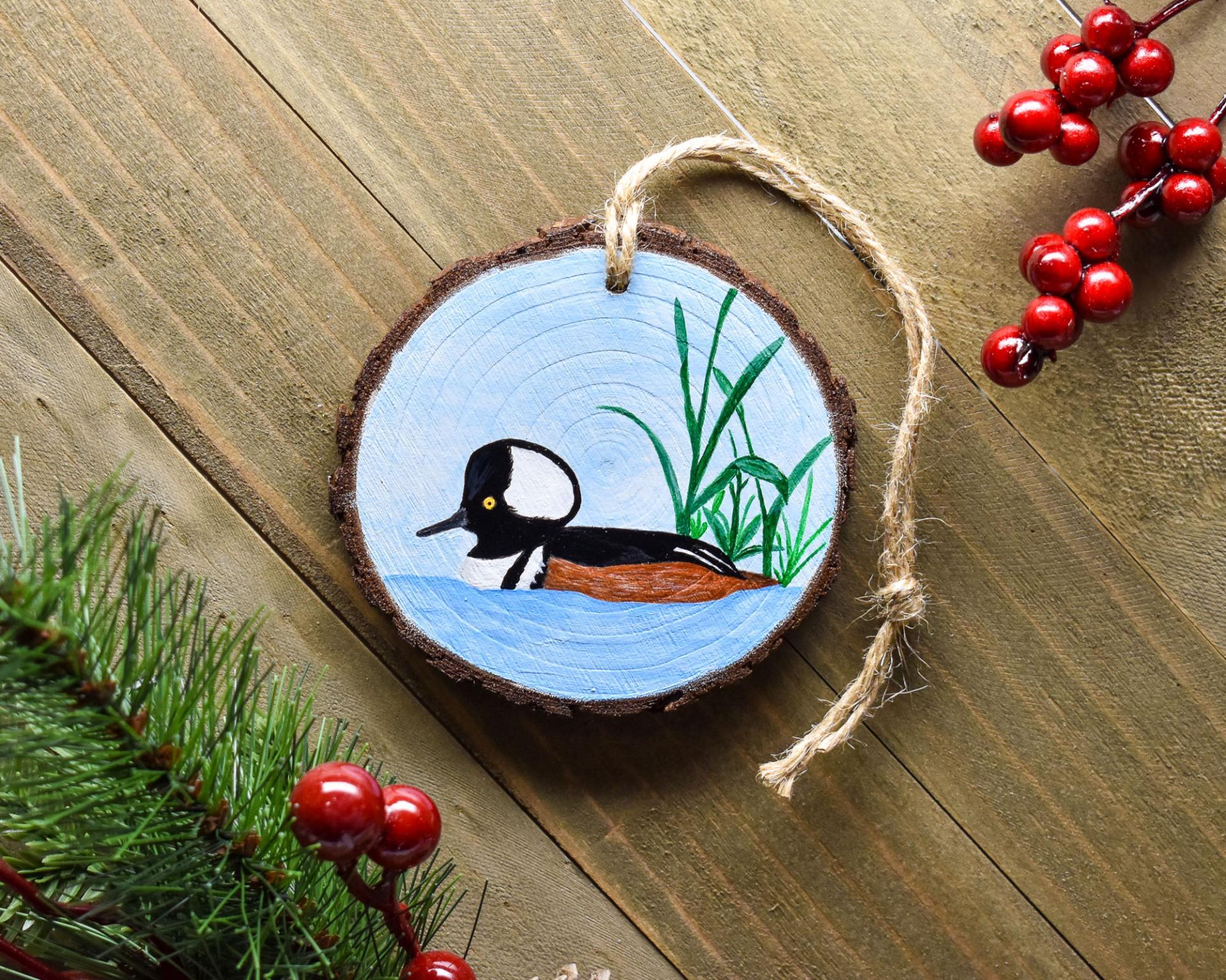 A handmade male hood merganser wood slice Christmas ornament displayed on a flat board at covecreekphotography.com.